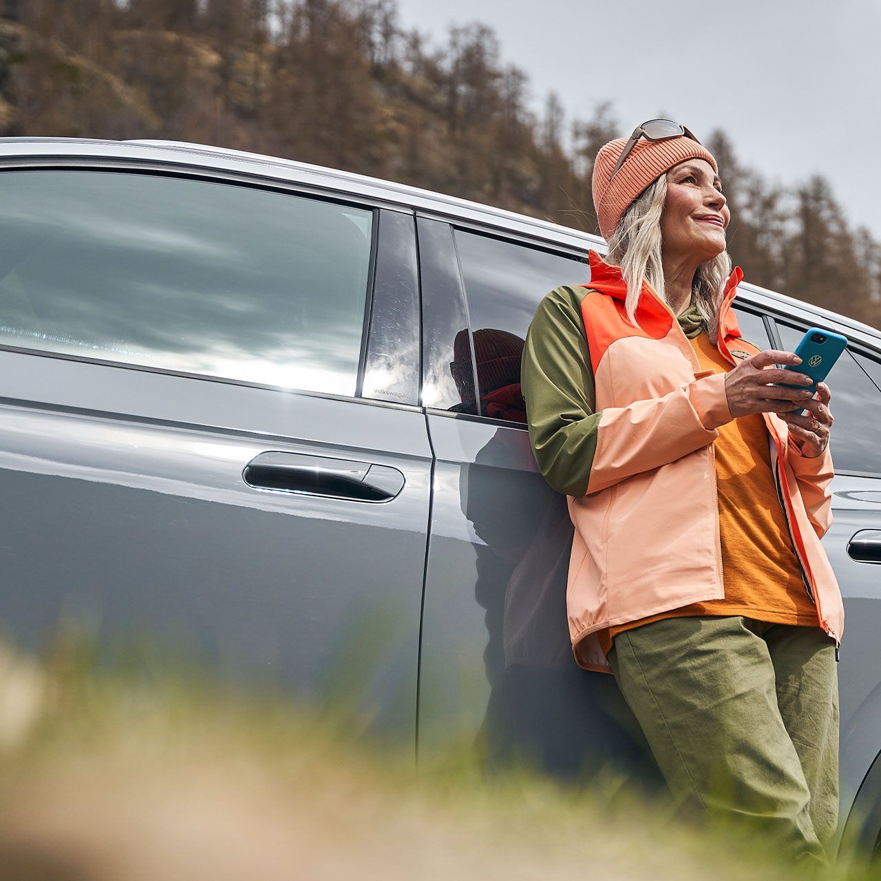 Eine Frau  mit Smartphone lehnt an einem Volkswagen und blickt in die Ferne