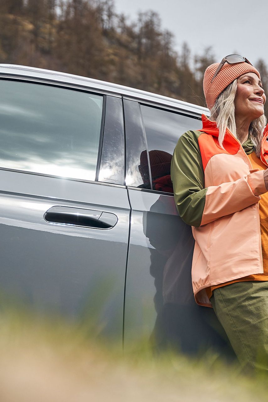 Eine Frau  mit Smartphone lehnt an einem Volkswagen und blickt in die Ferne