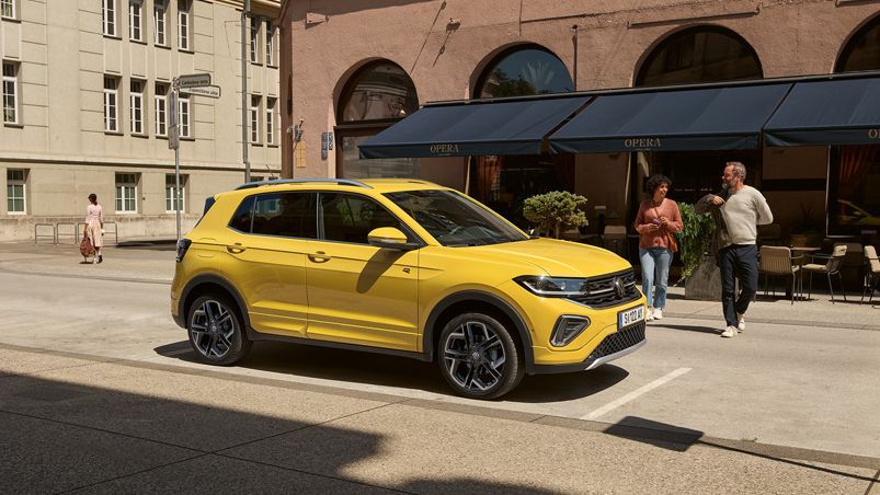 Ein gelber VW T-Cross parkt am Straßenrand in einer Stadt mit mehrstöckigen Gebäuden, ein Pärchen geht darauf zu.