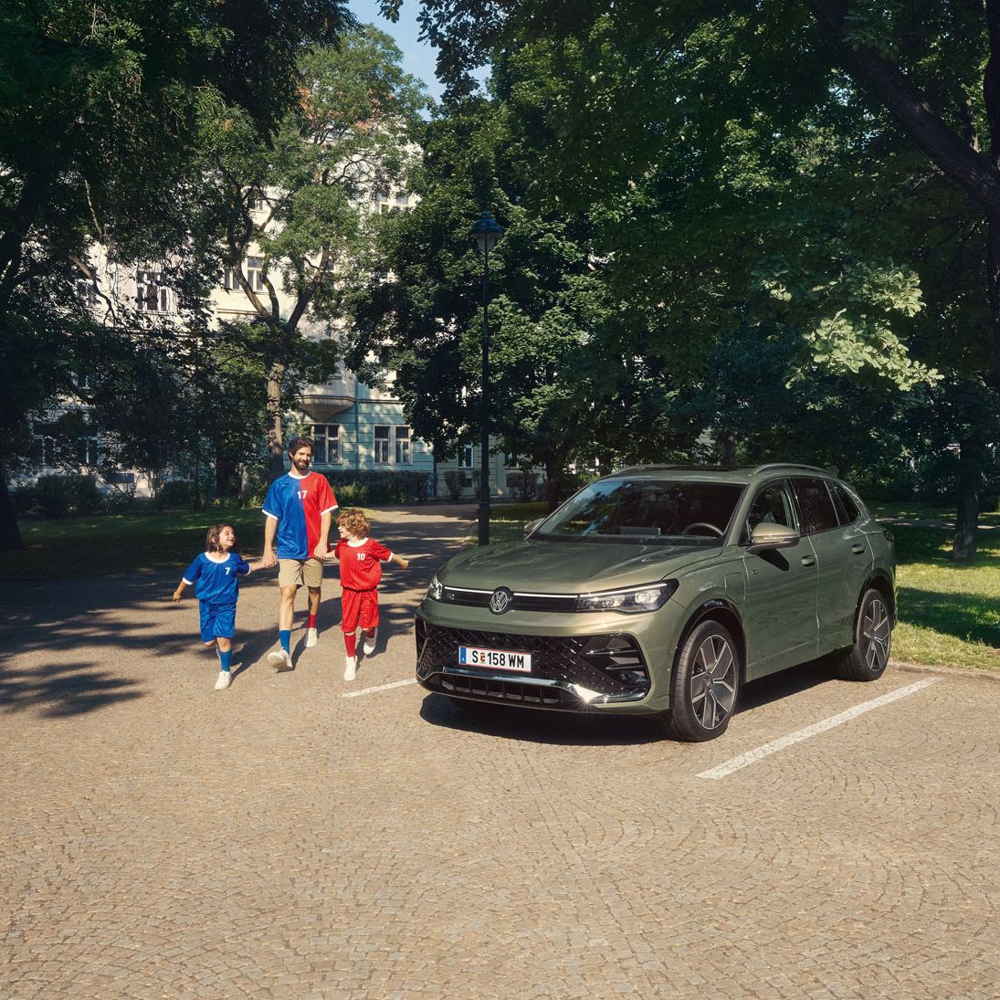 Ein Mann geht mit zwei Kindern von einem VW Tiguan R-Line Plug-In- Hybrid weg, der im Park steht.