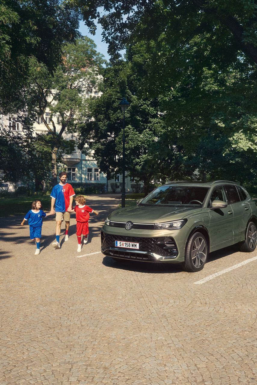 Ein Mann geht mit zwei Kindern von einem VW Tiguan R-Line Plug-In- Hybrid weg, der im Park steht.