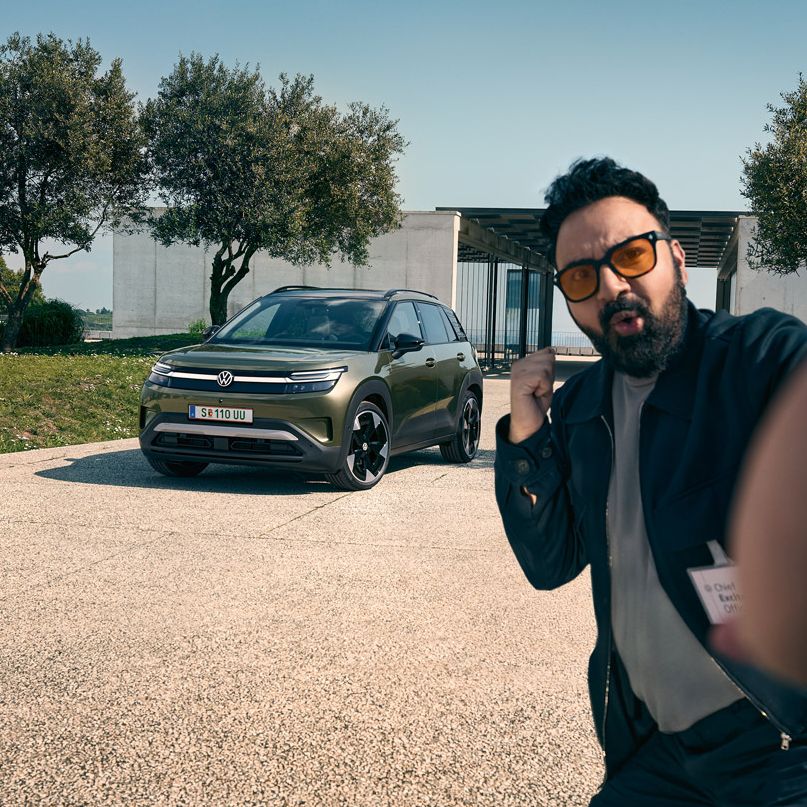 Ein Mann macht ein Selfie mit einem grünen VW ID. Cross, der auf der Auffahrt eines Gebäudes parkt.