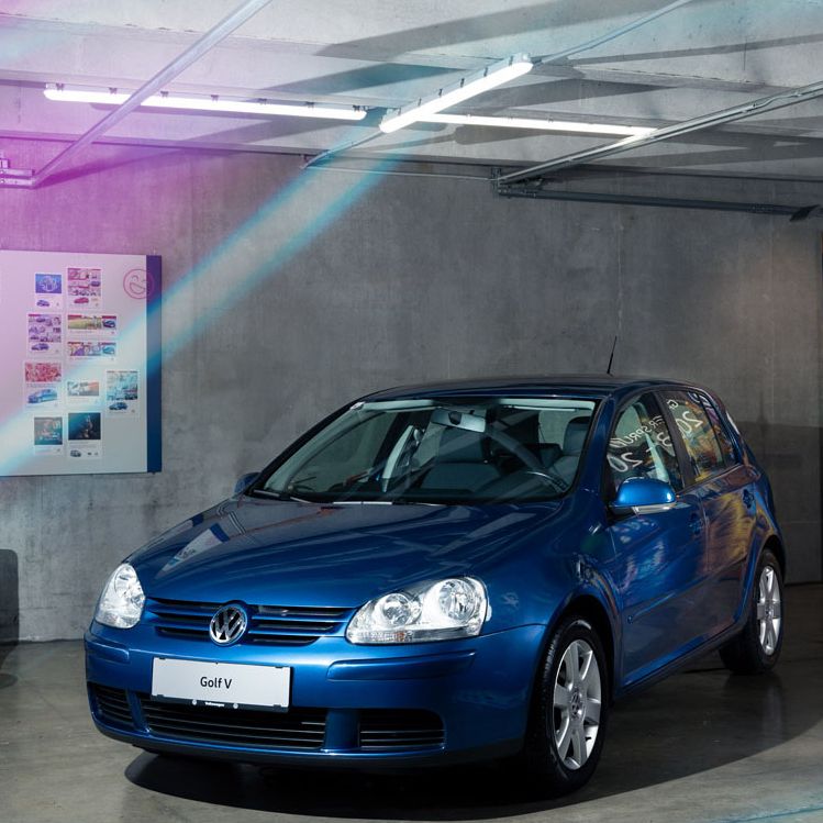 Ein blauer Volkswagen Golf V steht in einer modernen Garage. Im Hintergrund sind Wandtafeln mit Informationen und Bildern. Rechteckige Beleuchtungen und Neonlichter runden den futuristischen Stil ab.