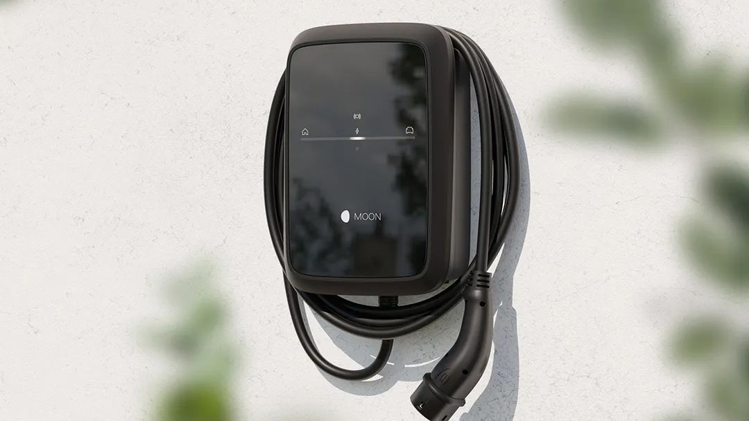 Schwarze Wallbox-Ladestation mit aufgewickeltem Ladekabel ist an einer hellen Hauswand montiert. Auf der glänzenden Front sind dezente Symbole und das Logo „MOON“ zu erkennen. Im Vordergrund sind unscharfe grüne Blätter zu sehen, die der Szene einen natürlichen Rahmen geben.