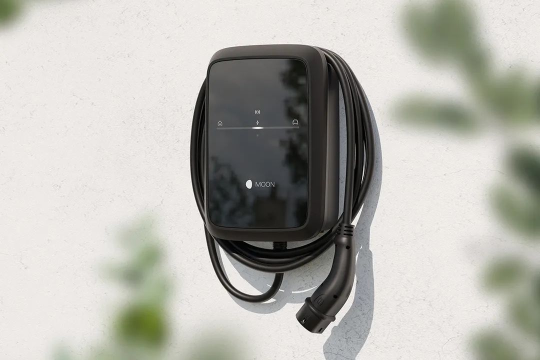 Schwarze Wallbox-Ladestation mit aufgewickeltem Ladekabel ist an einer hellen Hauswand montiert. Auf der glänzenden Front sind dezente Symbole und das Logo „MOON“ zu erkennen. Im Vordergrund sind unscharfe grüne Blätter zu sehen, die der Szene einen natürlichen Rahmen geben.