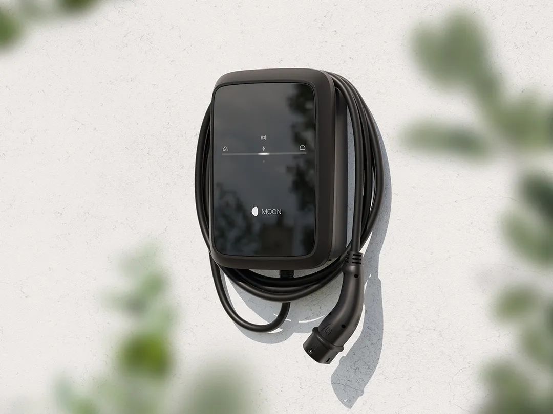 Schwarze Wallbox-Ladestation mit aufgewickeltem Ladekabel ist an einer hellen Hauswand montiert. Auf der glänzenden Front sind dezente Symbole und das Logo „MOON“ zu erkennen. Im Vordergrund sind unscharfe grüne Blätter zu sehen, die der Szene einen natürlichen Rahmen geben.