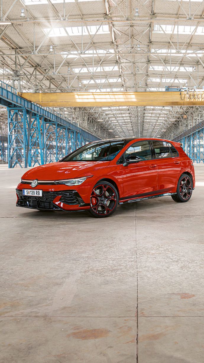 Ein roter Golf GTI steht in einer großen, leeren Industriehalle mit blauen Stahlträgern. Das Dach der Halle ist aus Glas und lässt viel Licht herein.