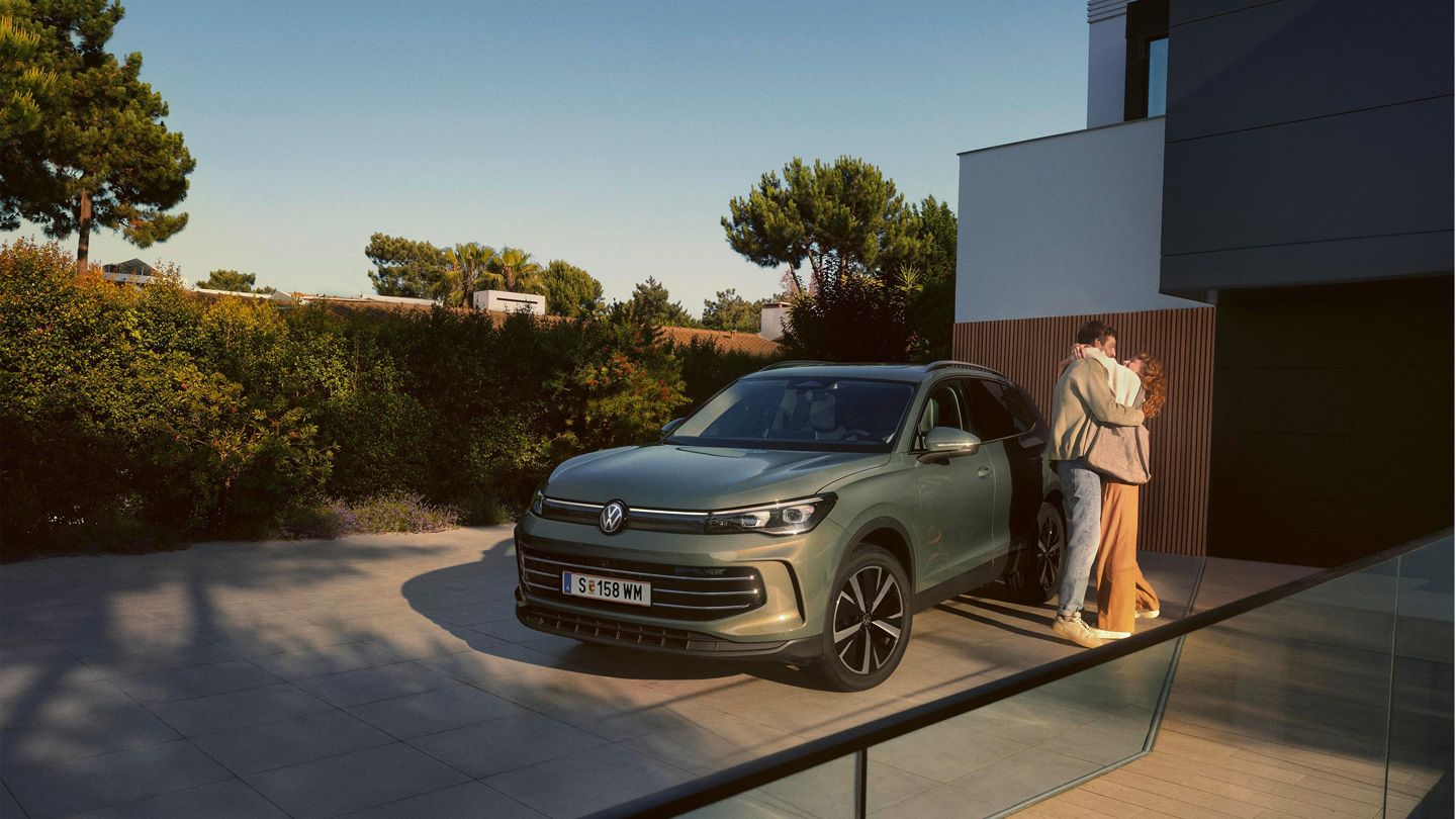 Eine Frau und ein Mann umarmen sich, neben ihnen steht ein VW Tiguan Elegance in der Farbe Cipressino-Grün Metallic