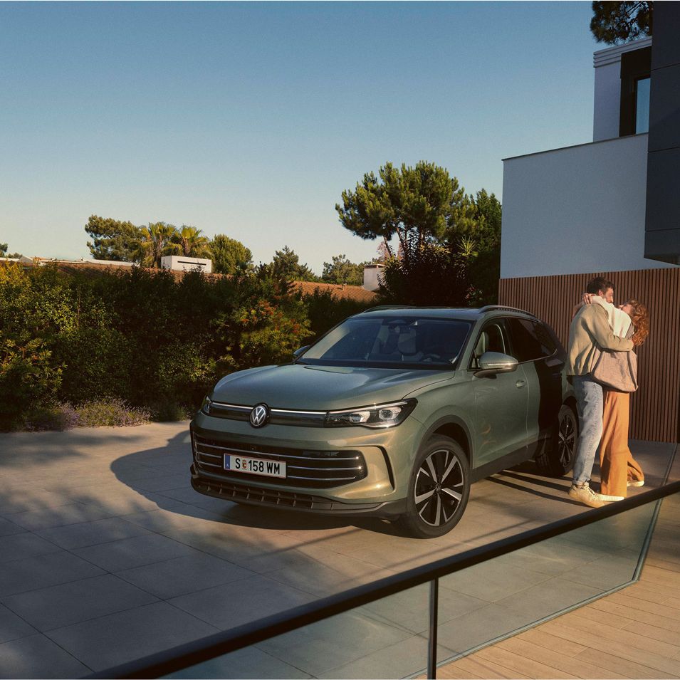 Eine Frau und ein Mann umarmen sich, neben ihnen steht ein VW Tiguan Elegance in der Farbe Cipressino-Grün Metallic