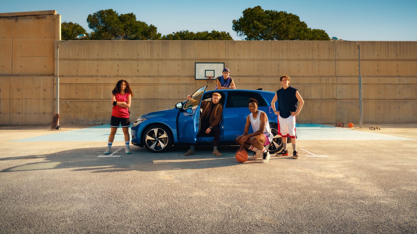 Fünf junge Erwachsene stehen und sitzen auf einem Basketballplatz neben einem blauen Auto; zwei halten Basketbälle in der Hand. Im Hintergrund ist ein Basketballkorb vor einer hohen Betonwand zu sehen, dahinter Bäume und ein klarer blauer Himmel. Die Szene wirkt entspannt und zeigt eine Mischung aus Sport- und Freizeitatmosphäre.
