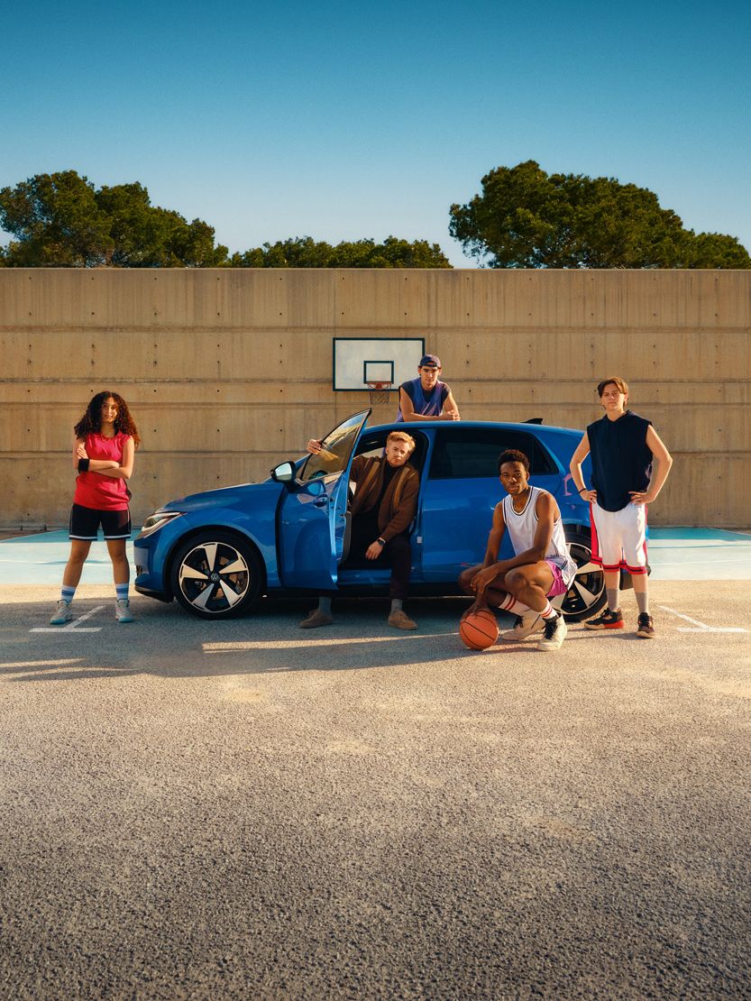 Fünf junge Erwachsene stehen und sitzen auf einem Basketballplatz neben einem blauen Auto; zwei halten Basketbälle in der Hand. Im Hintergrund ist ein Basketballkorb vor einer hohen Betonwand zu sehen, dahinter Bäume und ein klarer blauer Himmel. Die Szene wirkt entspannt und zeigt eine Mischung aus Sport- und Freizeitatmosphäre.