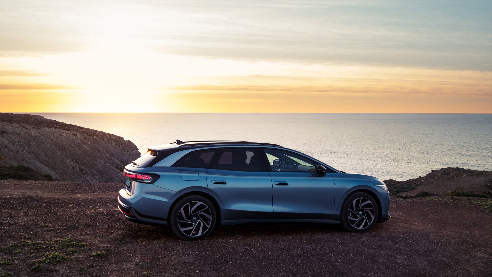 Ein blauer VW ID.7 Tourer steht oben auf einer Klippe im Sonnenuntergang, dahinter das Meer.