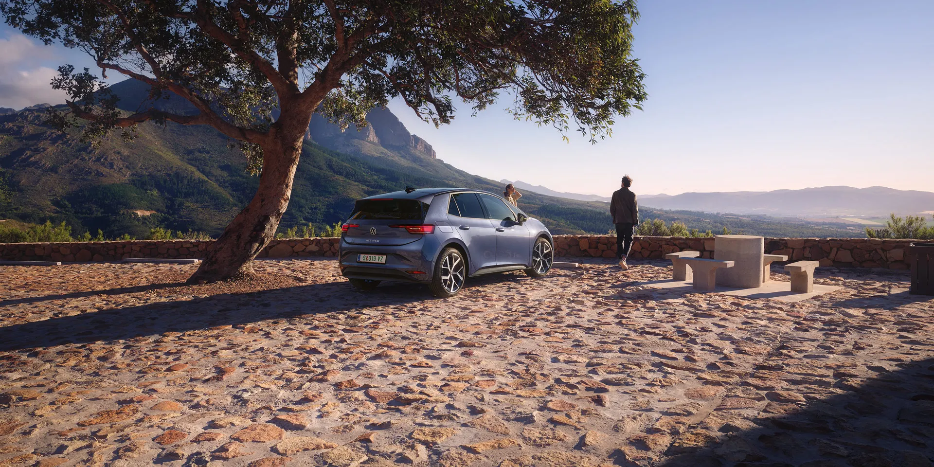 Auf einem Parkplatz in den Bergen parkt ein blauer VW ID.3 Neo, eine Frau und ein Mann genießen die Aussicht