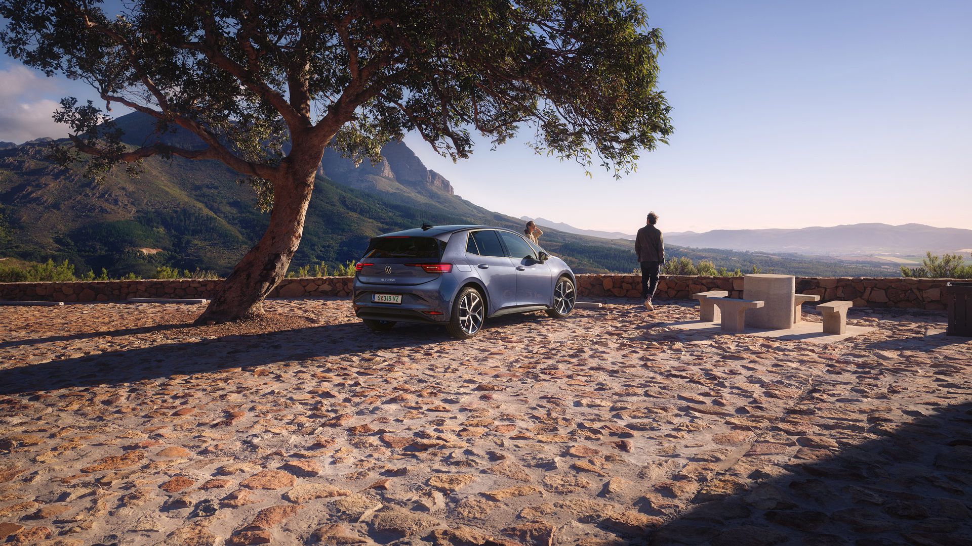 Auf einem Parkplatz in den Bergen parkt ein blauer VW ID.3 Neo, eine Frau und ein Mann genießen die Aussicht