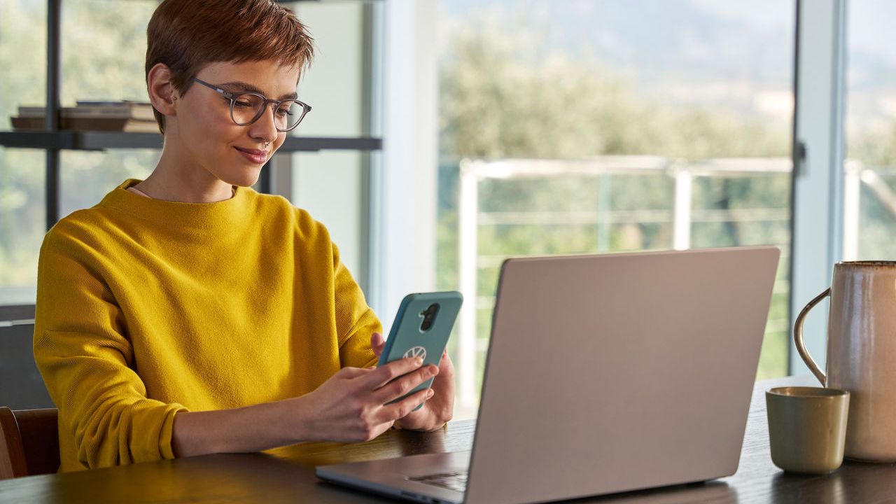 Eine Frau schaut vor ihrem Laptop auf Ihr Handy.