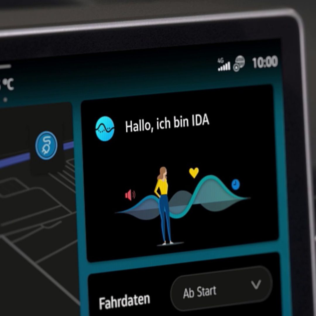 Ein Automonitor zeigt eine Begrüßungsmeldung "Hallo, ich bin IDA" mit einer stilisierten Figur vor grafischen Wellen. Oben rechts sind Uhrzeit und Signalstärke zu sehen. Darunter befindet sich ein Button mit der Aufschrift "Ab Start".