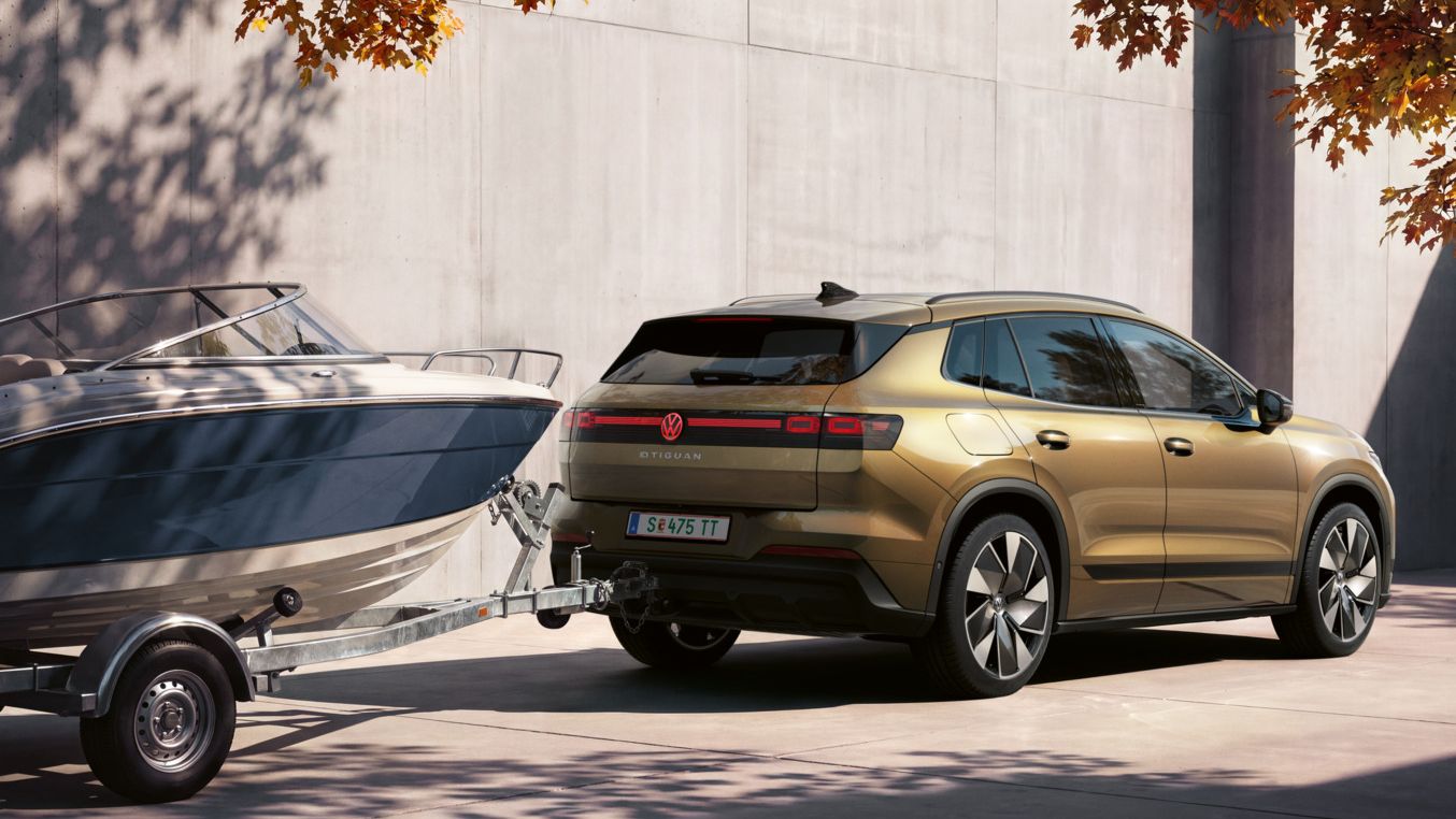 Ein oliv-bronzefarbener VW ID.4 Reskin parkt vor einem modernen Gebäude. Er zieht einen Anhänger mit Boot.