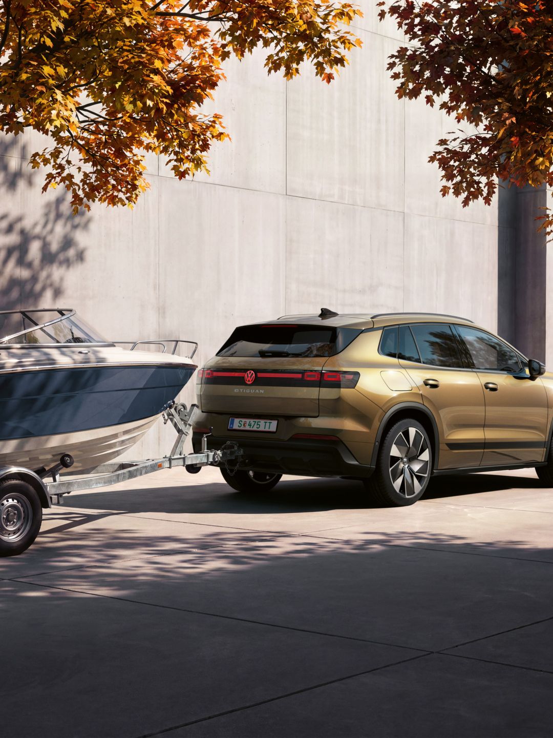 Ein oliv-bronzefarbener VW ID.4 Reskin parkt vor einem modernen Gebäude. Er zieht einen Anhänger mit Boot.