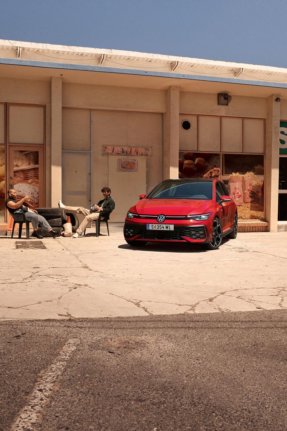 Der VW Golf GTI geparkt vor Gebäude, zwei junge Leute sitzen daneben in Stühlen