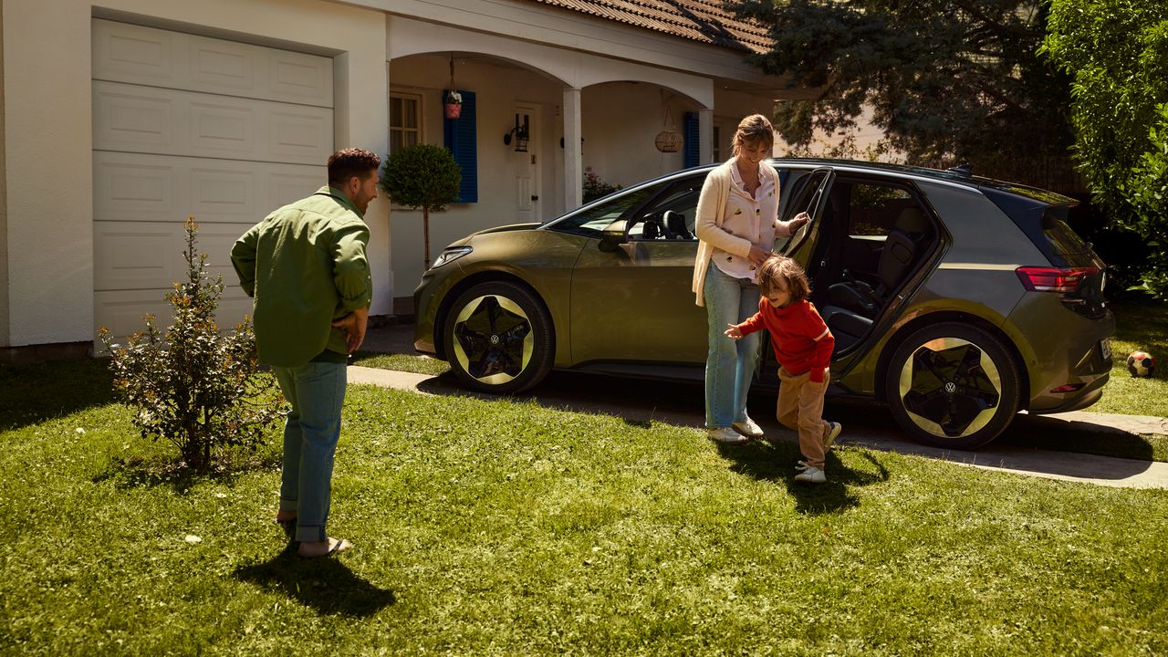 Ein ID.3 steht vor einem Wohnhaus. Die Mutter öffnet die hintere Tür, während ein Kind zum Vater auf den Rasen läuft
