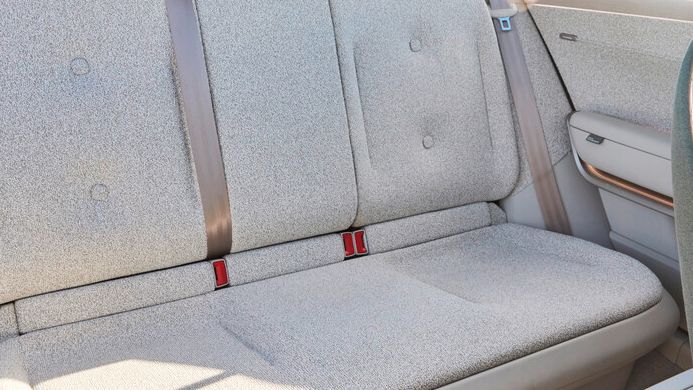 Bild eines Rücksitzes in einem Auto. Die Sitze sind mit einem hellen Stoff bezogen und haben zwei Sicherheitsgurte. Durch das Fenster sieht man eine Strandpromenade.