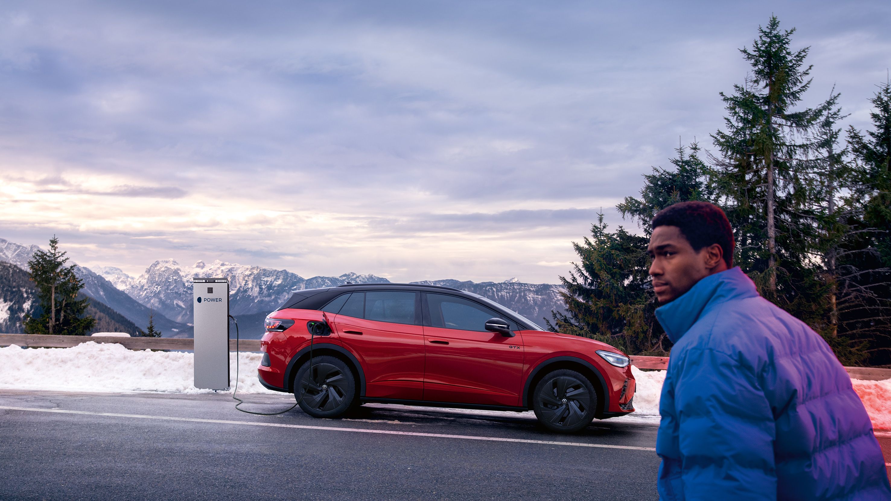 Seitenansicht eines roten VW ID.4 GTX der vor Bergkulisse geladen wird, Mann geht auf ihn zu