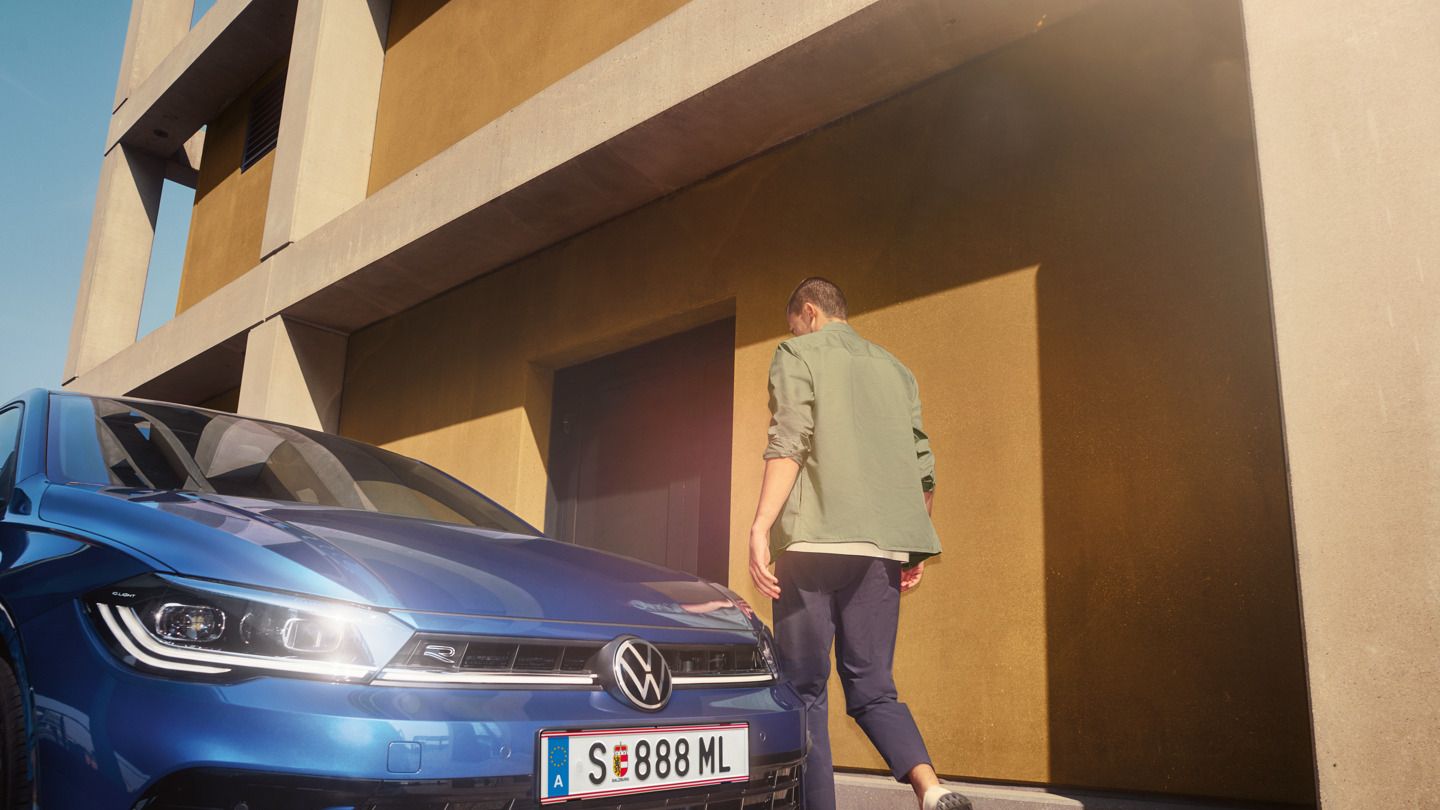 Ein Mann geht zur Fahrertür eines blauen VW Polo, der vor einem gelben Gebäude steht.