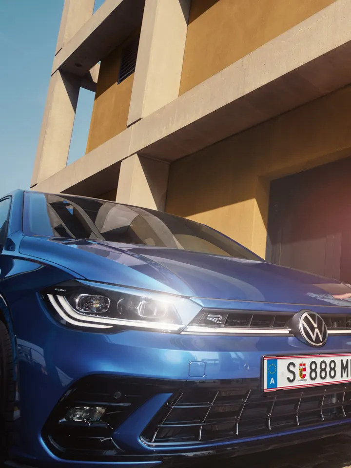 Ein Mann geht zur Fahrertür eines blauen VW Polo, der vor einem gelben Gebäude steht.