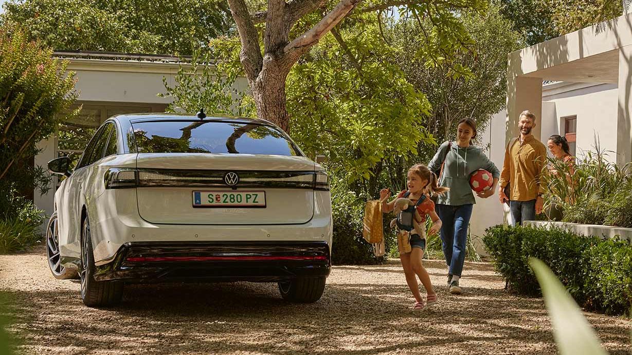 Eine Familie kommt gemeinsam mit einem Hund auf einem Grundstück an. Im Vordergrund sieht man ein Elektroauto, das in der Einfahrt geparkt ist. Die Umgebung ist grün und gut gepflegt.