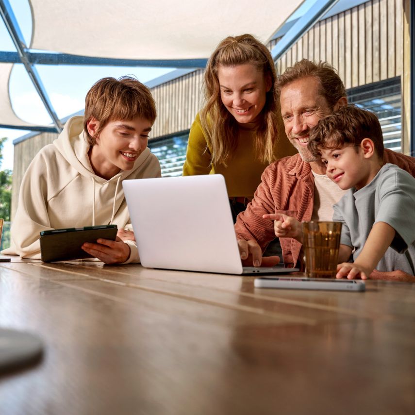 Eine Familie ist um einen Tisch versammelt und schaut fröhlich auf einen Laptop-Bildschirm