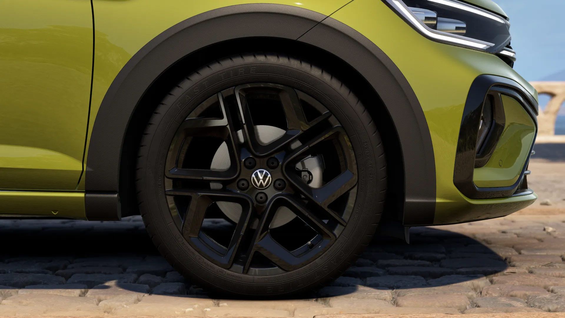 Nahaufnahme eines Autoreifens mit einem schwarzen Rad aus Metall und dem Volkswagen-Logo in der Mitte. Der Wagen ist olivgrün und steht auf einem gepflasterten Boden im Freien.