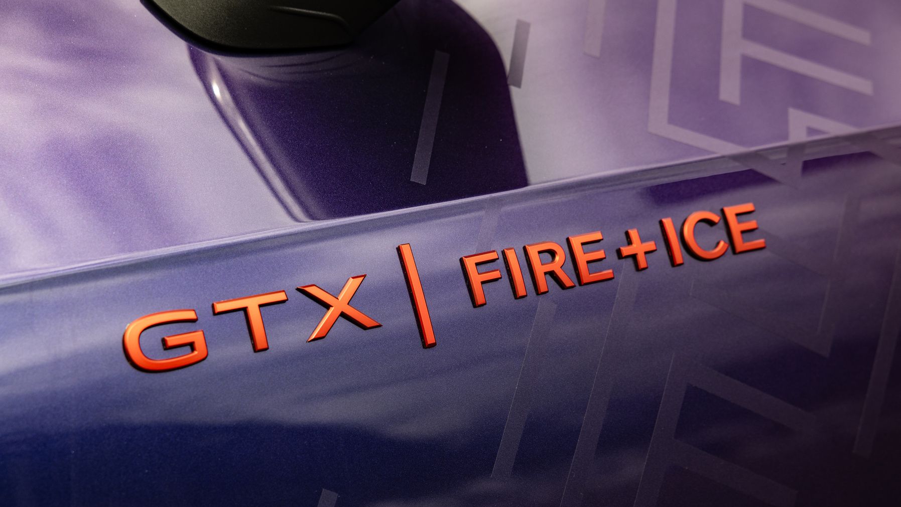 Nahaufnahme des Emblems "GTX FIRE+ICE" auf einem glänzenden violetten Fahrzeug. Das Emblem ist rot und hebt sich deutlich vom Fahrzeuglack ab. Hintergrund zeigt reflektiertes Licht und subtile Grafiken.