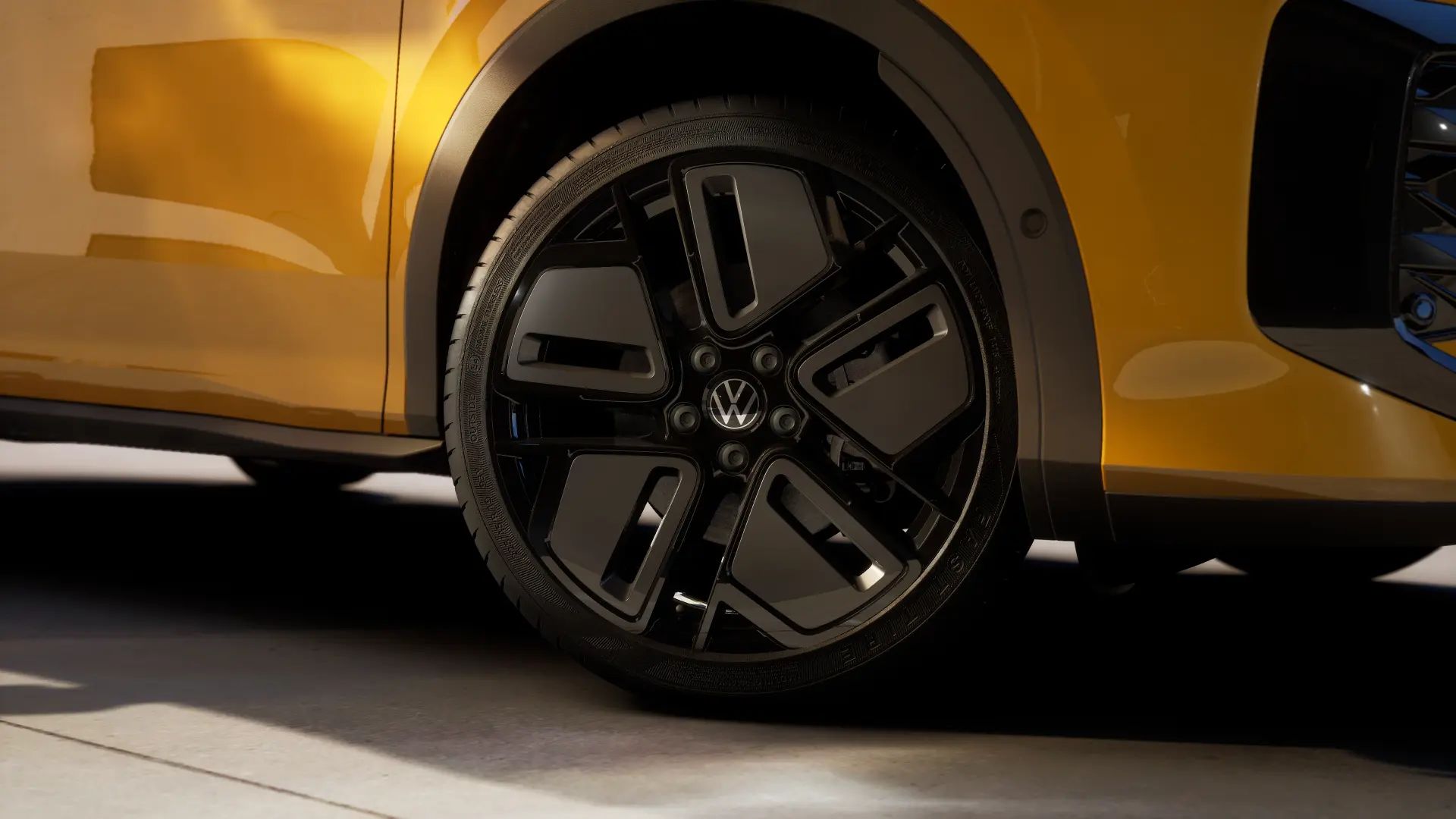 Nahaufnahme eines Autoreifens mit schwarzer Felge und Volkswagen-Logo, montiert an einem gelben T-Roc R-line. Der Reifen steht auf einem Betonboden. Das Fahrzeug hat eine moderne, glänzende Oberfläche.