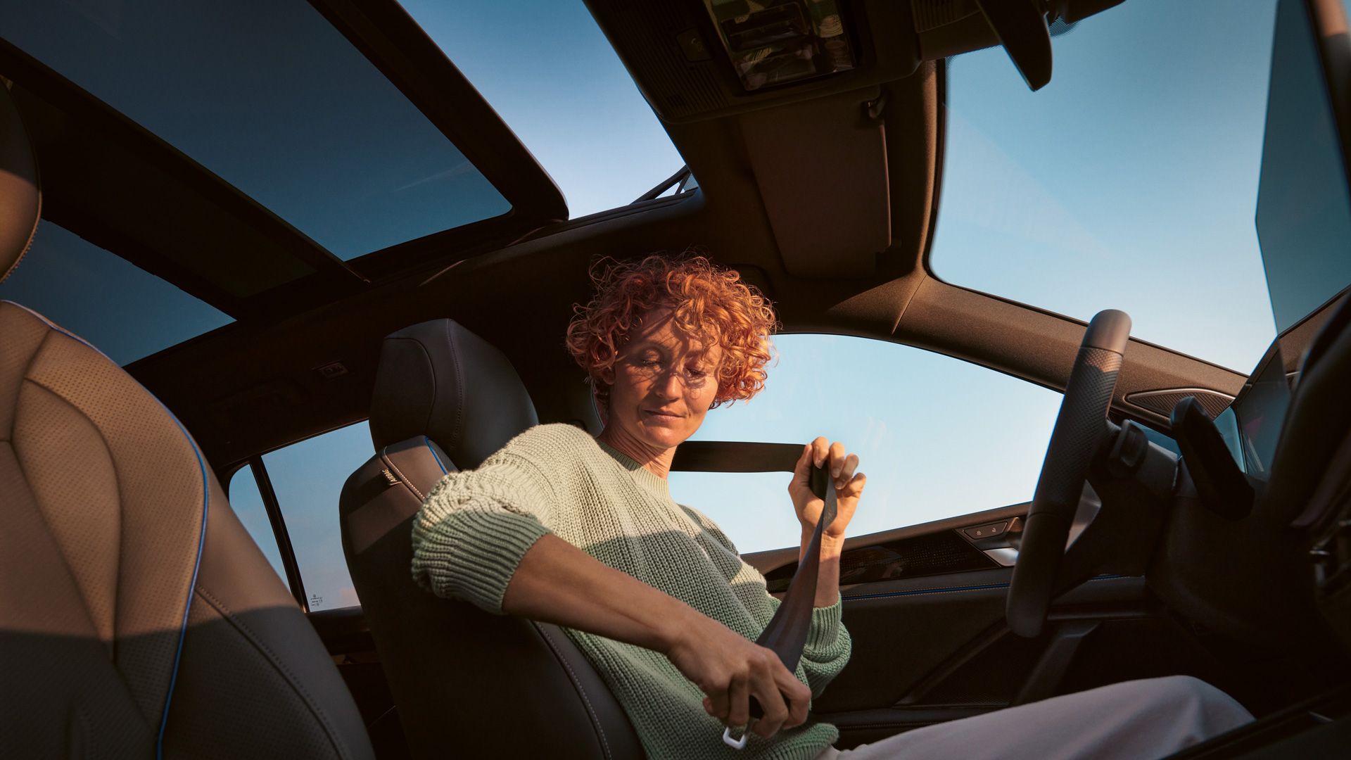 Eine Person mit lockigem Haar setzt im Innenraum eines Autos den Sicherheitsgurt an. Der Himmel draußen ist klar und blau. Das Gesicht der Person ist unscharf dargestellt.