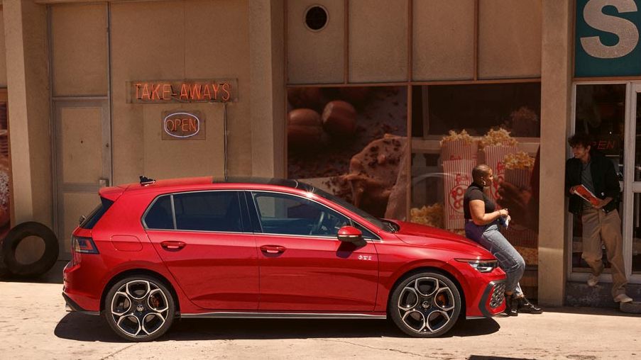 Roter VW Golf GTI steht vor einem Geschäft, eine Frau sitzt auf der Motorhaube.