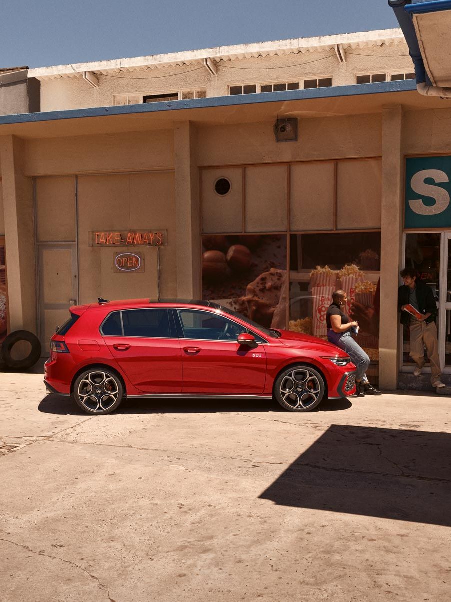 Roter VW Golf GTI steht vor einem Geschäft, eine Frau sitzt auf der Motorhaube.