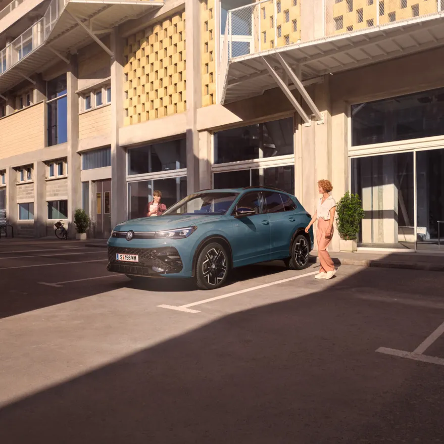 Eine junge Frau und ein junger Mann gehen zu ihrem roten Tiguan R-Line, der vor einem Bürogebäude auf einem Parkplatz steht.