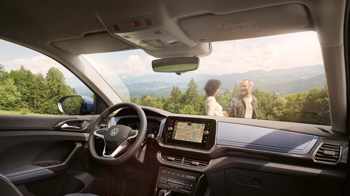 Blick vom Beifahrersitz eines blauen VW T-Cross durch die Frontscheibe auf ein lachendes Pärchen vor Wald und Bergen.