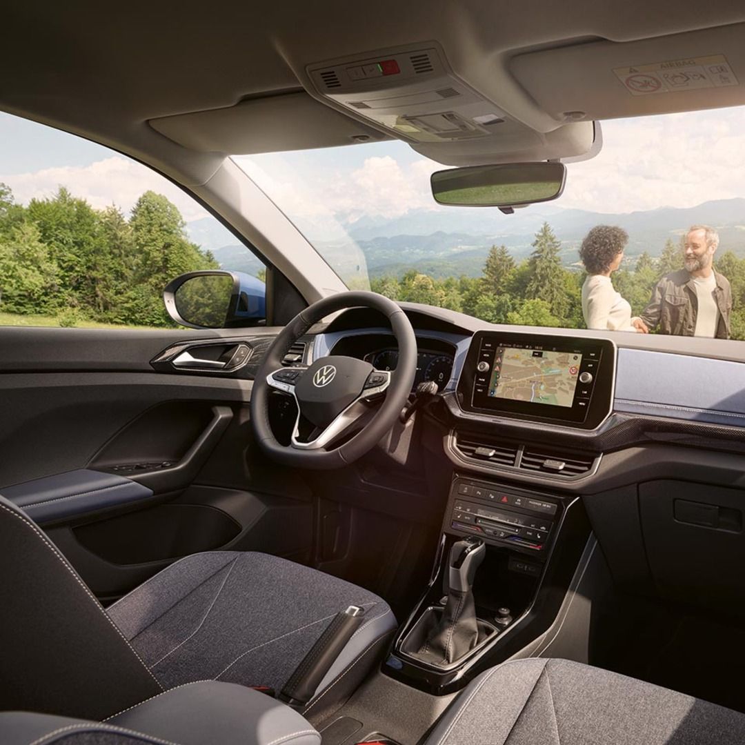 Blick vom Beifahrersitz eines blauen VW T-Cross durch die Frontscheibe auf ein lachendes Pärchen vor Wald und Bergen.