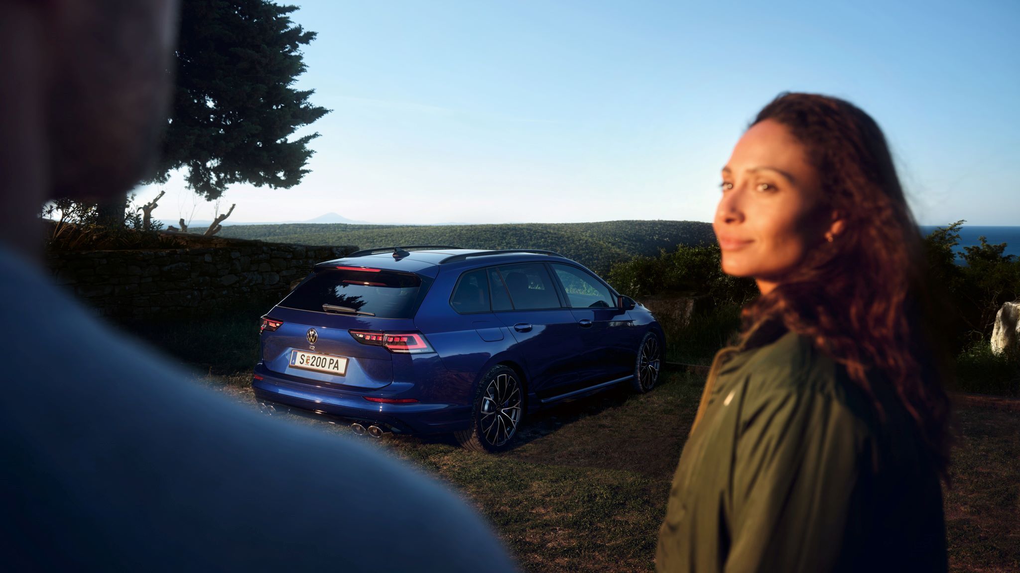 Frau und Mann im Vordergrund, im Hintergrund geparkter Golf Variant R in Heckansicht. 