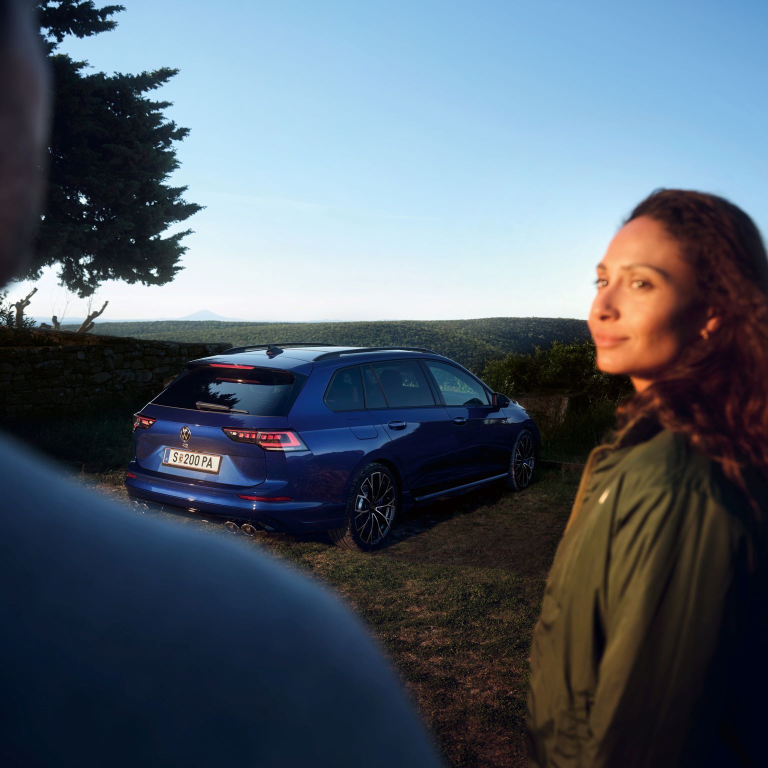 Frau und Mann im Vordergrund, im Hintergrund geparkter Golf Variant R in Heckansicht. 