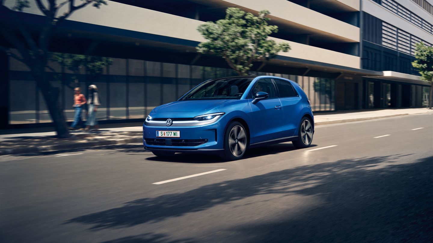 Ein blauer VW ID. Polo Style fährt durch eine Stadt mit grünen Bäumen am Straßenrand.