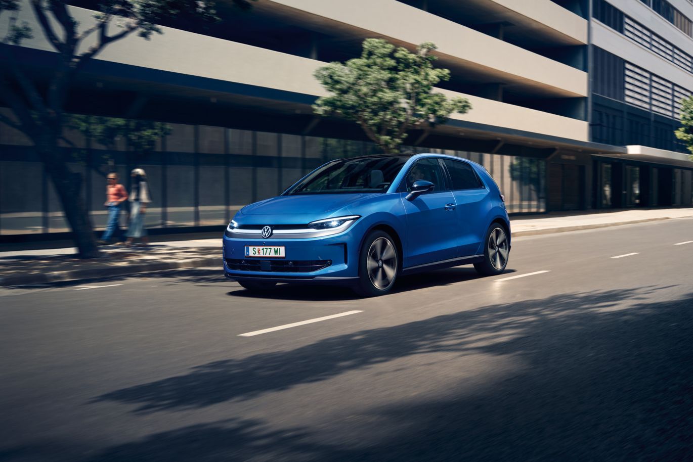 Ein blauer VW ID. Polo Style fährt durch eine Stadt mit grünen Bäumen am Straßenrand.