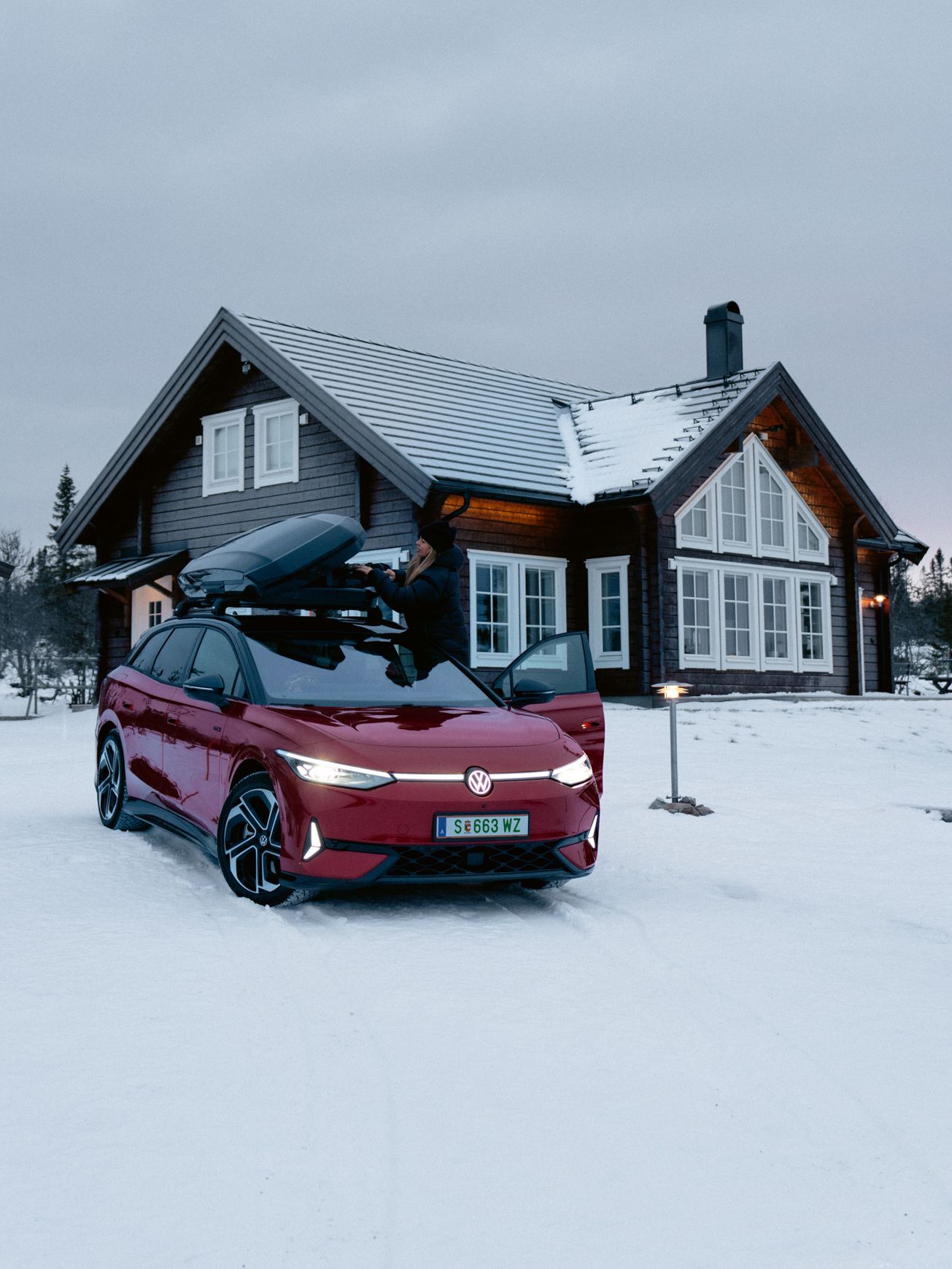 Ein roter ID.7 Tourer GTX mit Dachbox steht im Schnee vor einem Wohnhaus. 