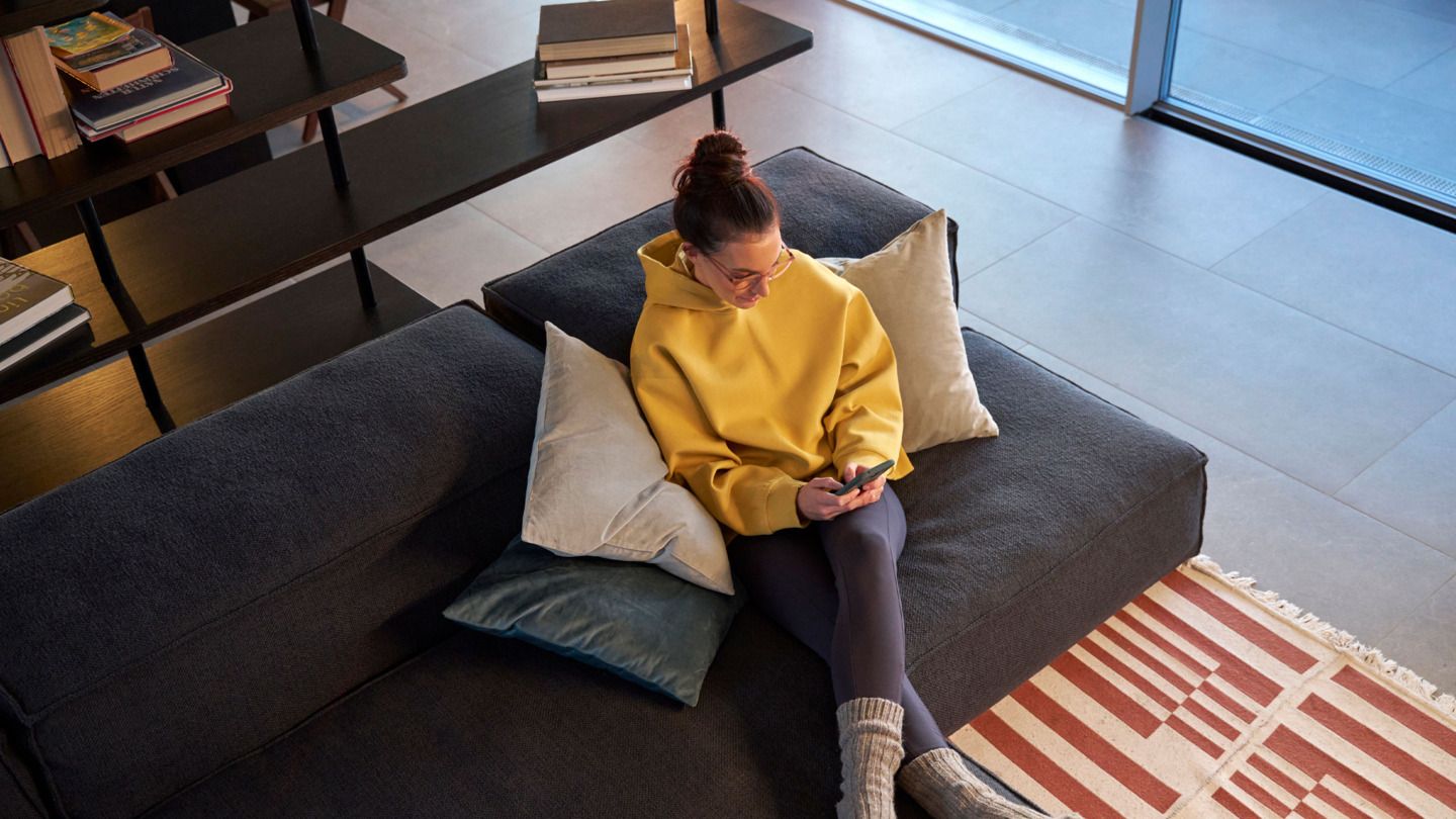 Person sitzt in einem modernen Wohnzimmer auf einem grauen Sofa und hält ein Handy in der Hand. Sie trägt einen gelben Hoodie, schwarze Leggings und graue Socken. Ein Bücherregal voller Bücher steht im Hintergrund.