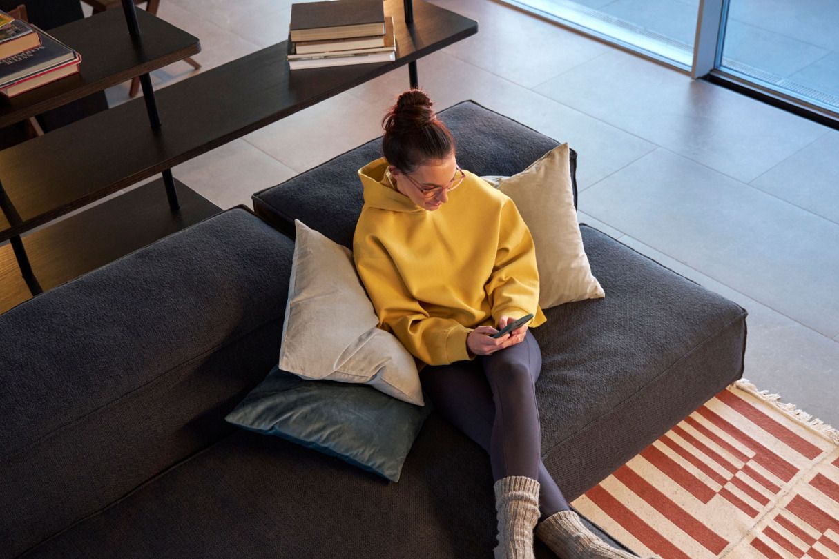 Person sitzt in einem modernen Wohnzimmer auf einem grauen Sofa und hält ein Handy in der Hand. Sie trägt einen gelben Hoodie, schwarze Leggings und graue Socken. Ein Bücherregal voller Bücher steht im Hintergrund.