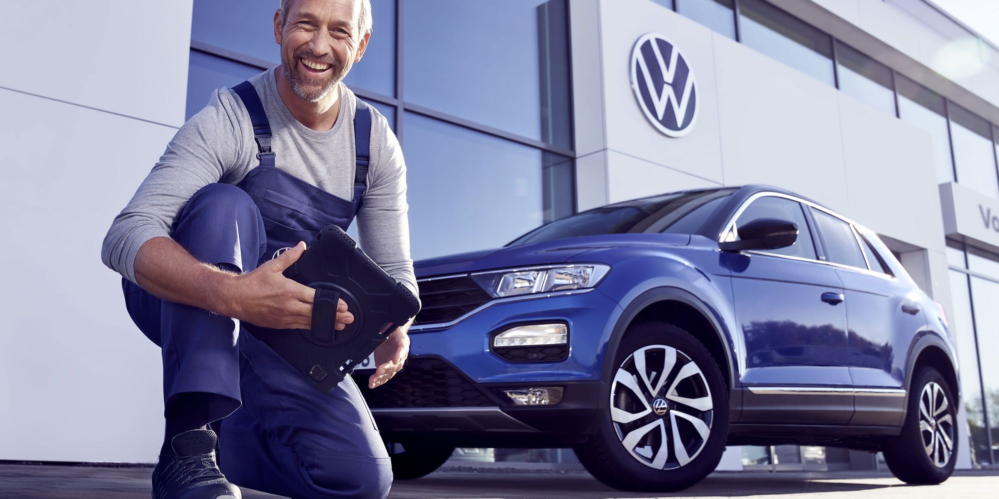 Ein Volkswagen Service Techniker mit Tablet, kniet vor einem blauen Volkswagen T-Roc