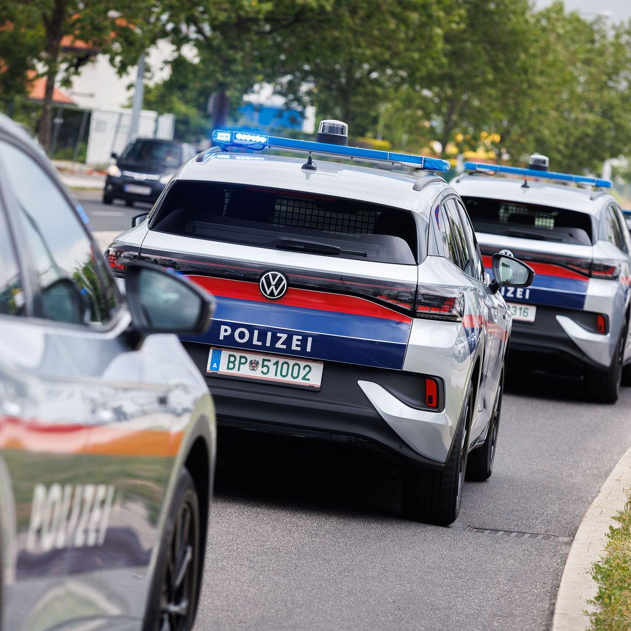 Eine Reihe von österreichischen Polizeiautos fährt auf einer Straße. Die Fahrzeuge haben Blaulichter und sind weiß mit blauen und roten Streifen. Im Hintergrund sind Bäume und Gebäude zu sehen.