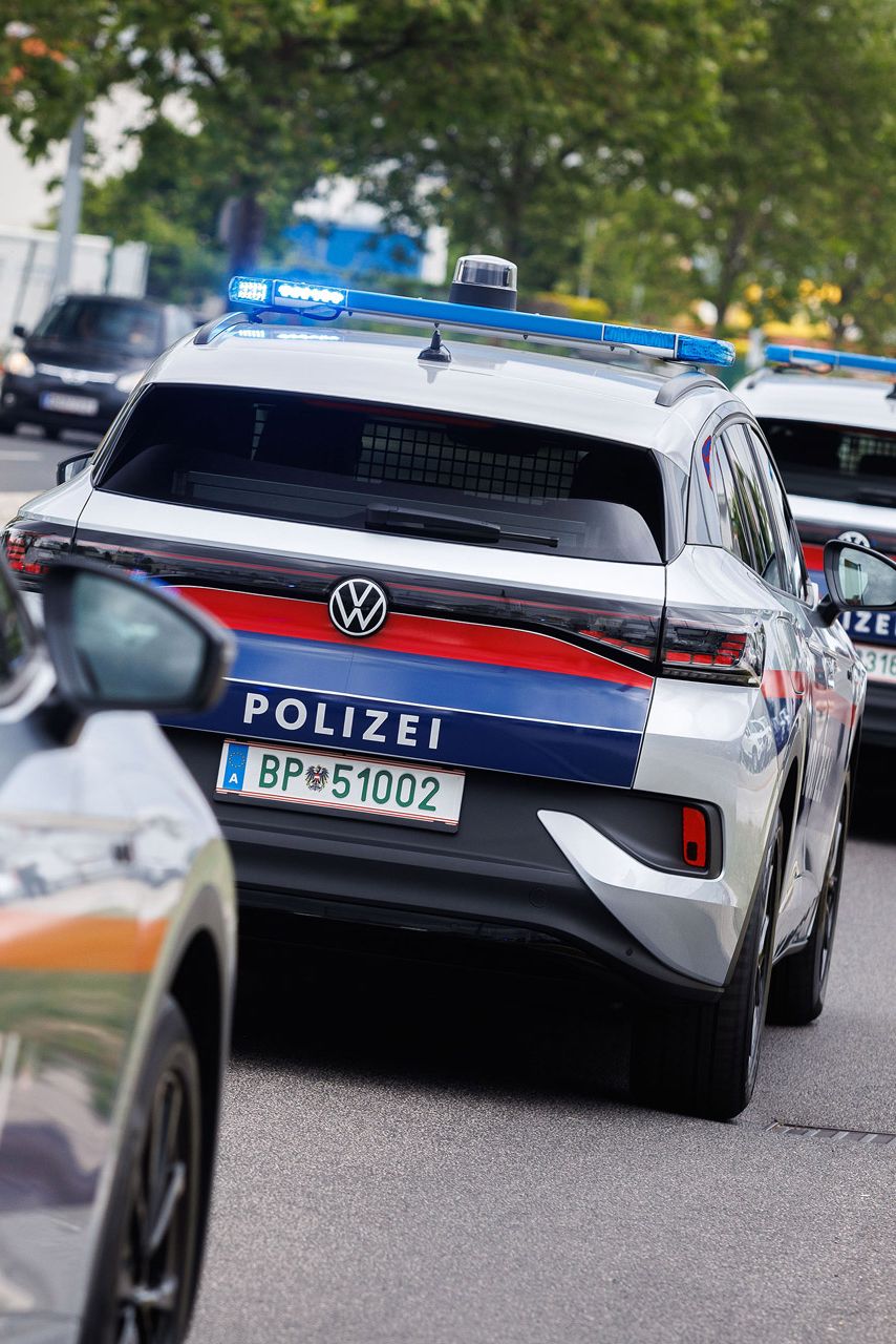 Eine Reihe von österreichischen Polizeiautos fährt auf einer Straße. Die Fahrzeuge haben Blaulichter und sind weiß mit blauen und roten Streifen. Im Hintergrund sind Bäume und Gebäude zu sehen.