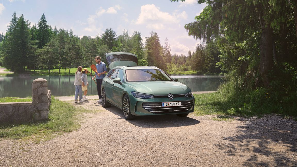Der neue VW Passat Elegance in Maripositgrün Metallic parkt an einem Badesee mit geöffnetem Kofferraum. Ein Mann verteilt Freitzeitspielzeug an zwei Kinder.